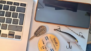A flat lay image showing a laptop keyboard, smartphone, fishing gear including a lure, hooks, line, and a weight, representing the crossover between technology and fishing.