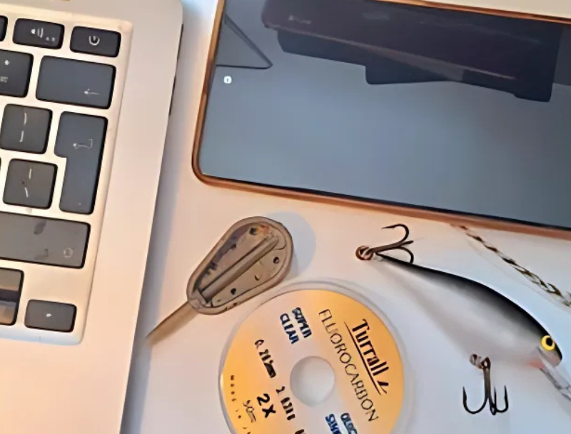A flat lay image showing a laptop keyboard, smartphone, fishing gear including a lure, hooks, line, and a weight, representing the crossover between technology and fishing.